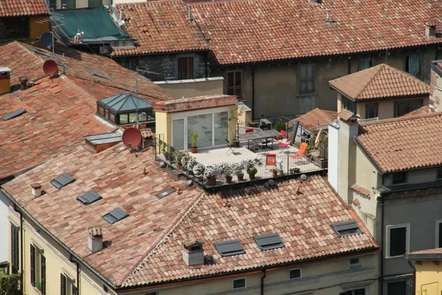 Tejados de tejas rojizas con ventanas, chimeneas y antenas, junto a una terraza con macetas, mesa y sillas.